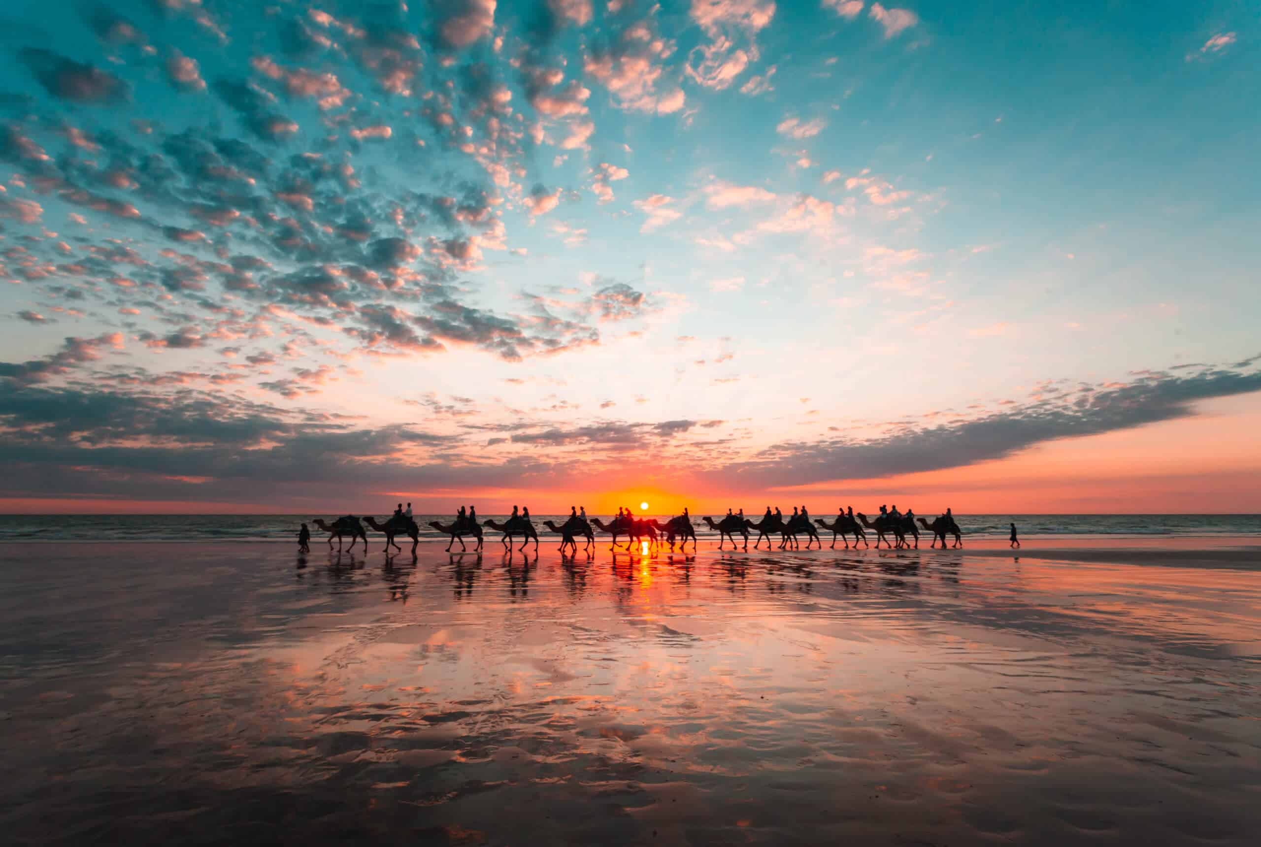 A photo of a beautiful beach in Broome, Western Australia, with the sun setting in the background and the silhouette of camels in the distance.
